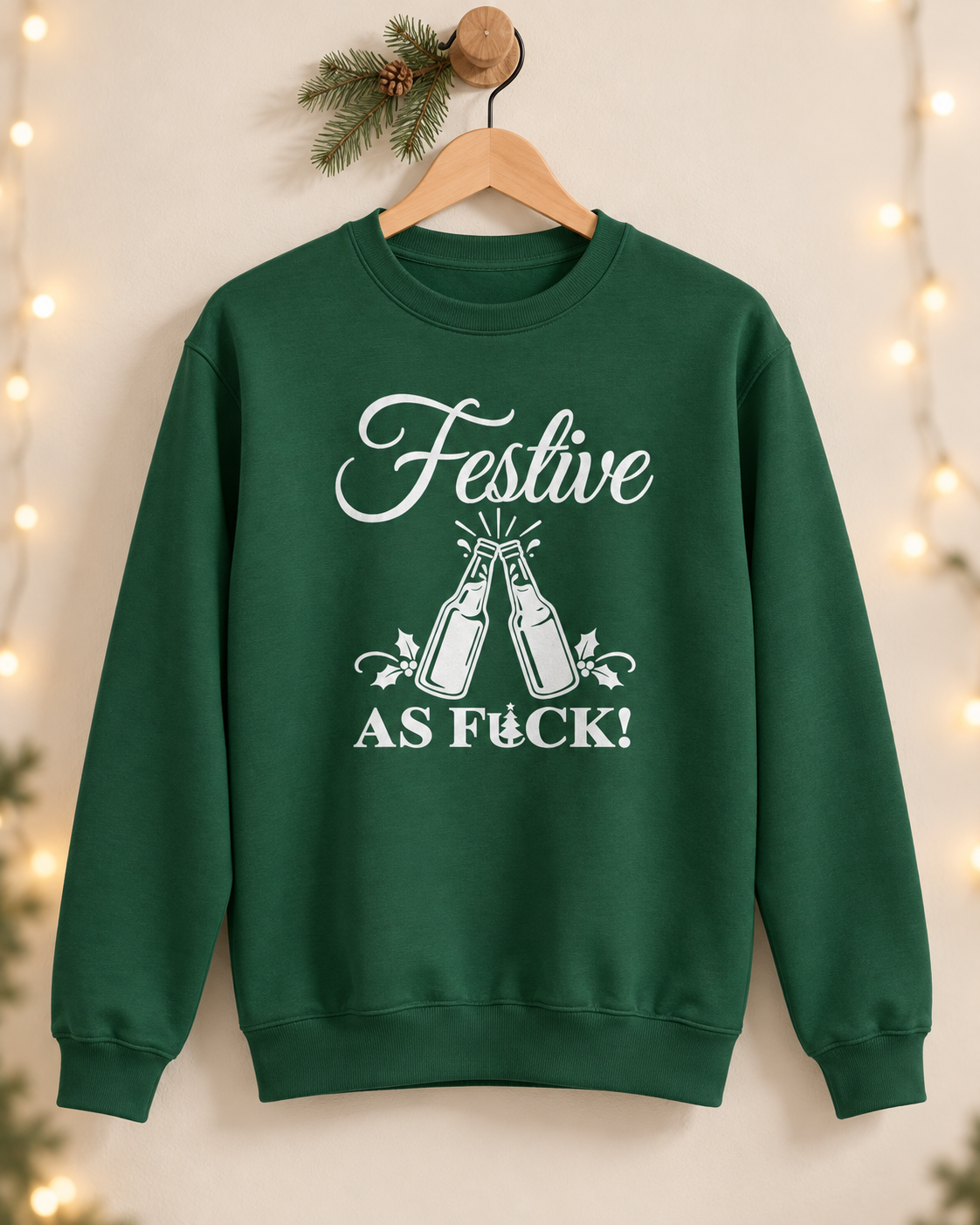 A dark green adult Christmas crewneck sweatshirt hanging on a wooden hanger against a neutral wall with string lights and a pine sprig, featuring a white graphic of clinking beer bottles and the text "FESTIVE AS FUCK!".