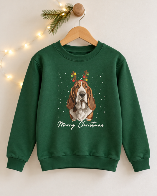 Dark green kids Christmas crewneck sweatshirt hanging on a wooden hanger, featuring a graphic of a Basset Hound dog wearing reindeer antlers with colourful baubles and 'Merry Christmas' script below.