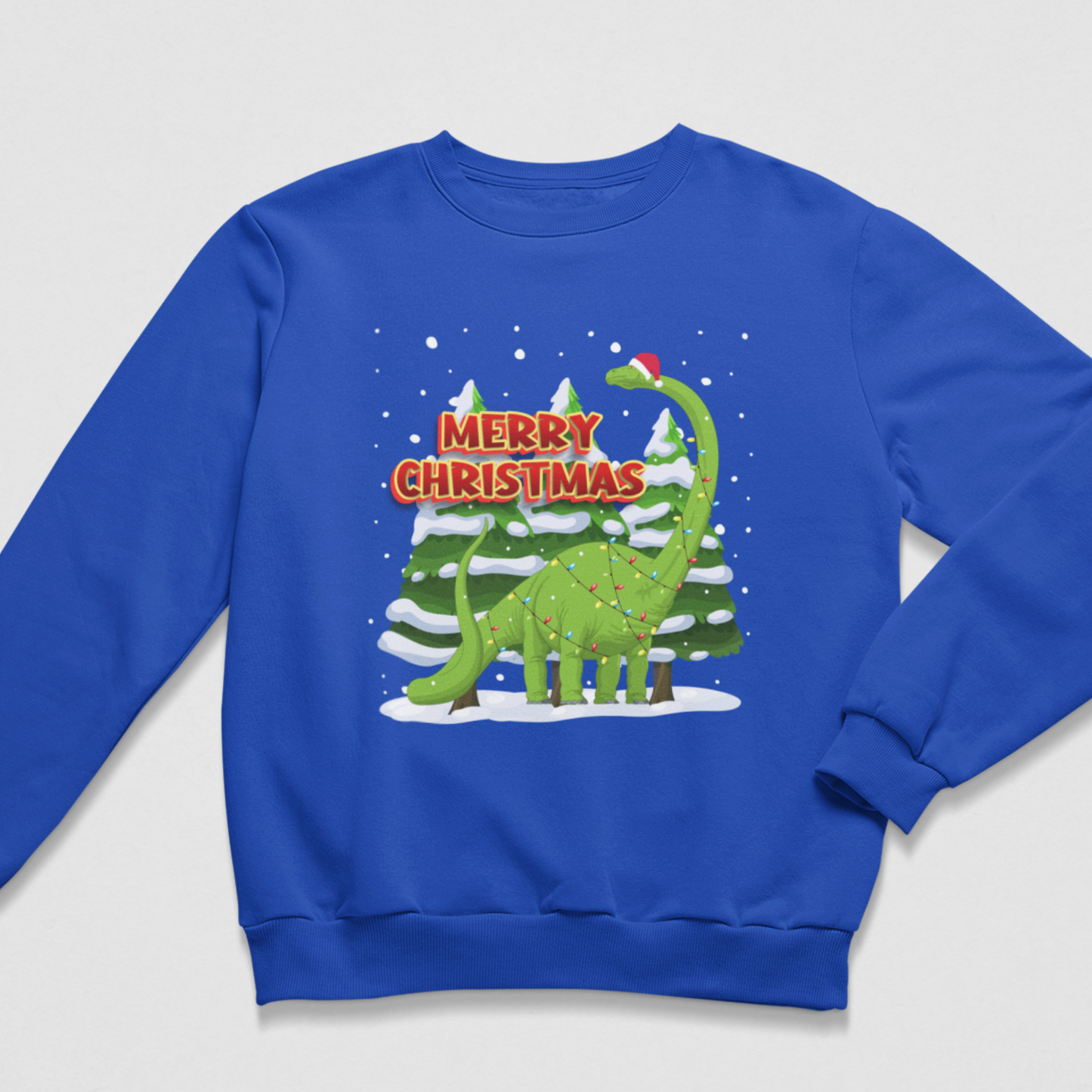 A kids royal blue crewneck sweatshirt featuring a festive Christmas graphic of a green long-necked dinosaur wearing a Santa hat, standing in front of snow-covered pine trees with 'MERRY CHRISTMAS' text in red and white.