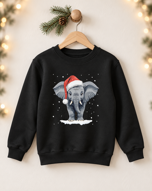 A kids black crewneck sweatshirt hanging on a wooden hanger against a warm beige wall decorated with soft holiday lights and pine branches. The sweatshirt features a graphic print of a grey elephant wearing a red Santa hat with white trim, standing in white snow against a dark background.