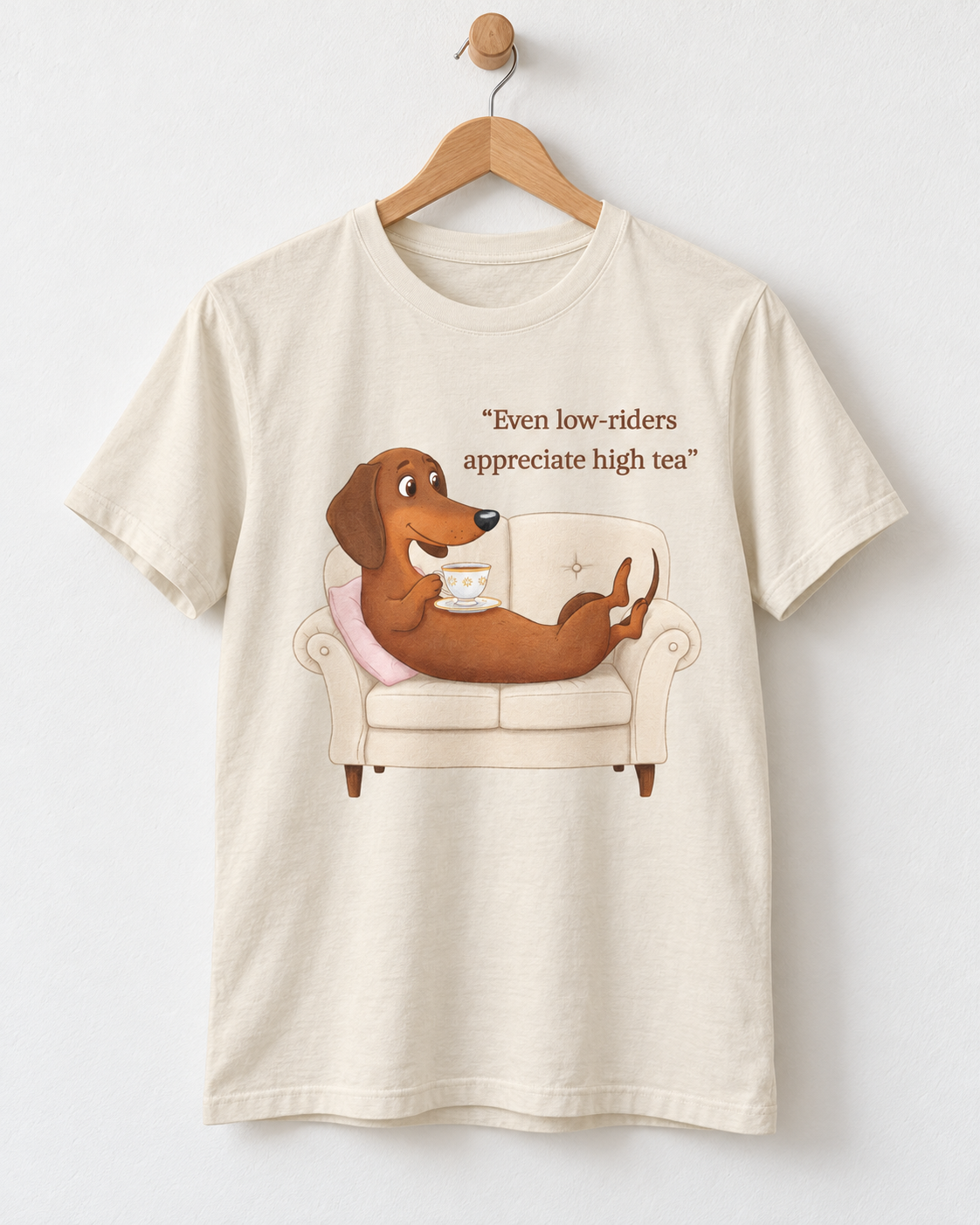 A cream-colored short-sleeve t-shirt hanging on a wooden hanger against a white wall. The shirt features a cartoon graphic of a brown dachshund sitting on a beige armchair holding a teacup, with the text 'Even low-riders appreciate high tea' printed above.
