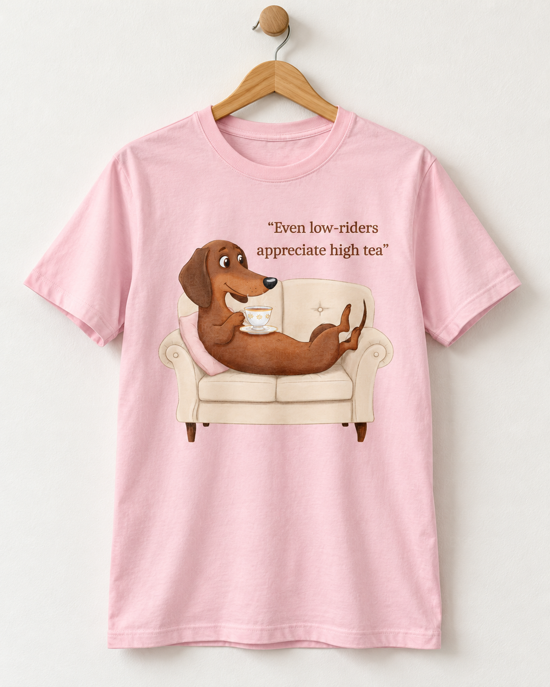 A light pink short-sleeve crewneck t-shirt hanging on a wooden hanger against a light gray wall. The shirt features a graphic of a brown dachshund relaxing on a cream-colored armchair, holding a teacup, with the text 'Even low-riders appreciate high tea' printed above.