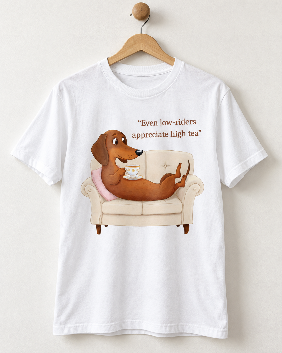 A white short-sleeve crewneck t-shirt hanging on a wooden hanger against a light beige wall. The shirt features a cartoon graphic of a brown dachshund sitting comfortably on a cream-colored armchair, holding a teacup, with the text 'Even low-riders appreciate high tea' printed above the illustration.