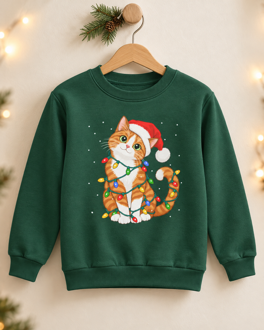 A dark green kids' Christmas crewneck sweatshirt hanging on a wooden hanger against a neutral wall with a pine sprig and string lights, featuring an illustration of an orange and white tabby cat wearing a Santa hat and tangled in colourful festive fairy lights.