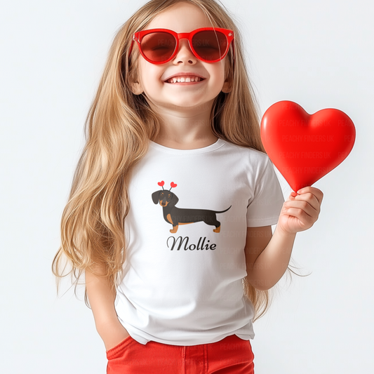 Smiling girl wearing a personalised white t-shirt with a sausage dog graphic and red heart antenna headband, with the name “Mollie” printed underneath.