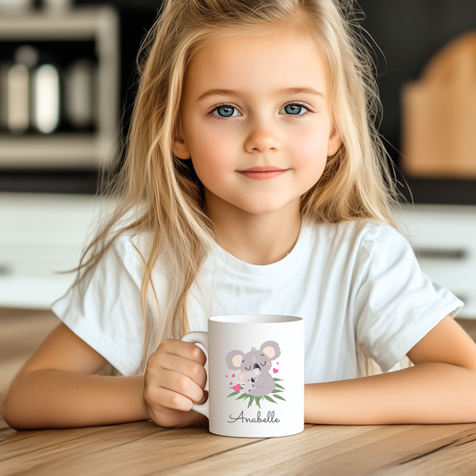 Young girl sitting at a table holding a white personalised mug featuring a mother and baby koala design with the name “Anabelle” printed underneath.