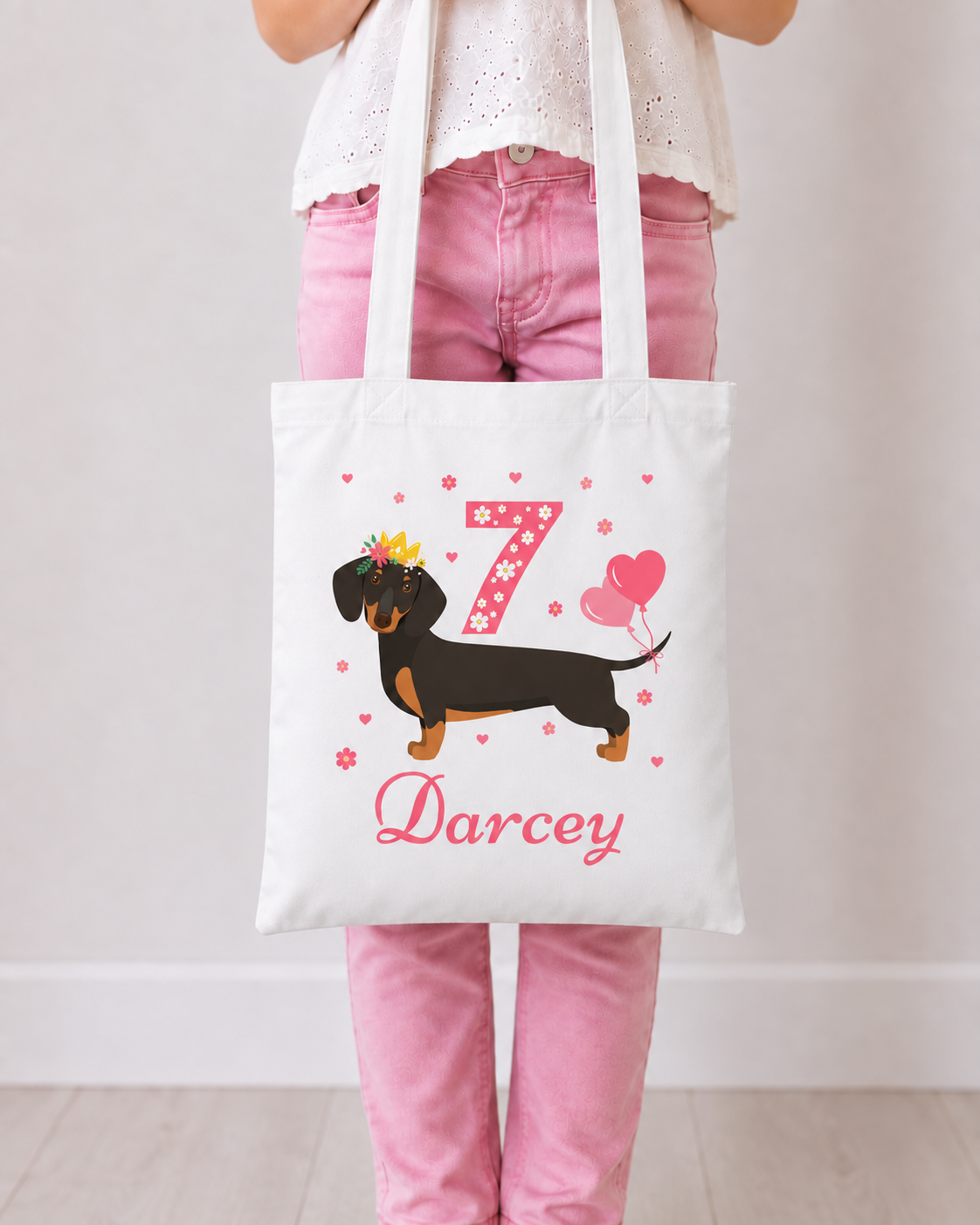 Girl in pink jeans holding a white personalised tote featuring a crowned dachshund, pink floral number birthday number 7, heart balloons and the name Darcey.