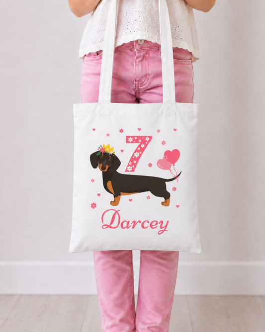 Girl in pink jeans holding a white personalised tote featuring a crowned dachshund, pink floral number birthday number 7, heart balloons and the name Darcey.