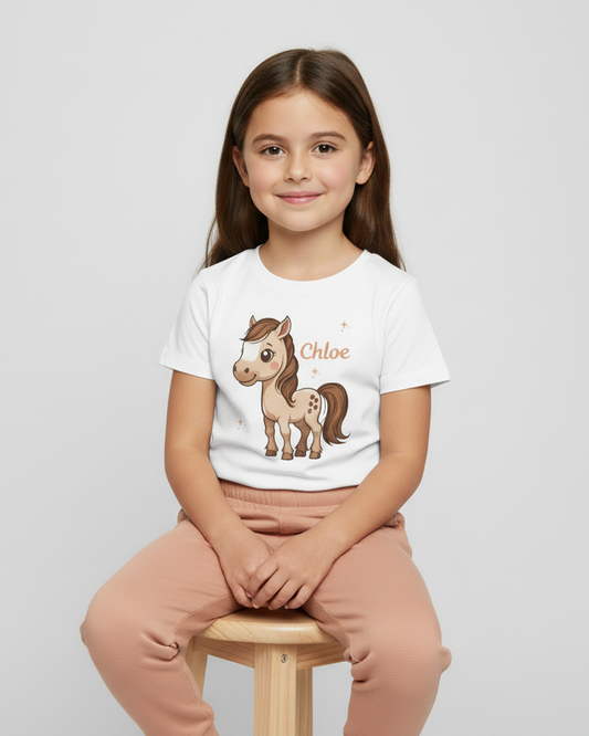 Girl sitting on a wooden stool wearing a custom name pony T-shirt in white with dusty peach joggers, photographed on a light grey studio backdrop.