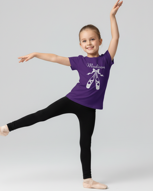 Side view of a young girl in a purple personalised ballet t-shirt with ballet shoes design, black leggings, and ballet flats, holding a graceful dance pose with arms extended in a light studio setting.