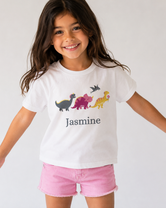 Young girl models a white personalised dinosaur t-shirt featuring colourful cartoon dinosaurs walking in a line and the name "Jasmine" printed below.