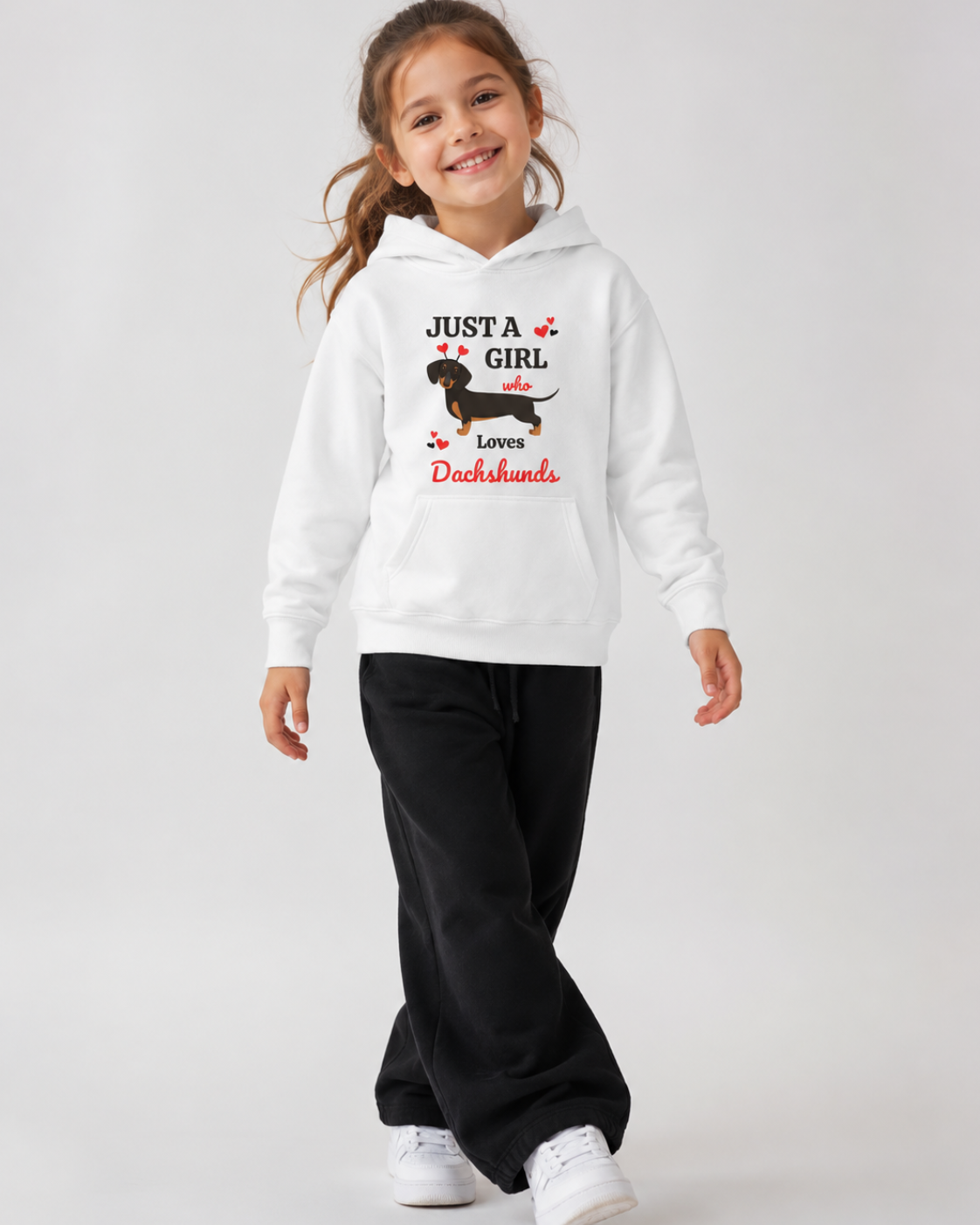 Smiling girl in a white “Just a Girl Who Loves Dachshunds” hoodie with a black and tan sausage dog graphic, styled with wide-leg black joggers in a relaxed mid-step studio pose.