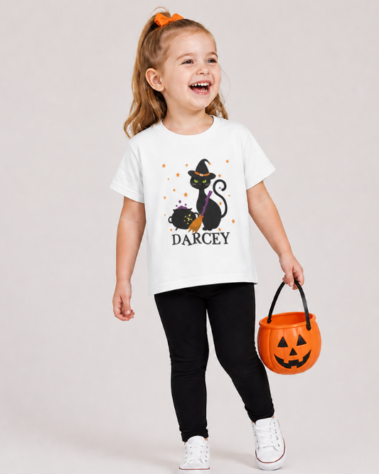 Little girl wearing personalised Halloween cat t-shirt with name Darcey, kids white graphic tee with black leggings holding pumpkin bucket, studio background