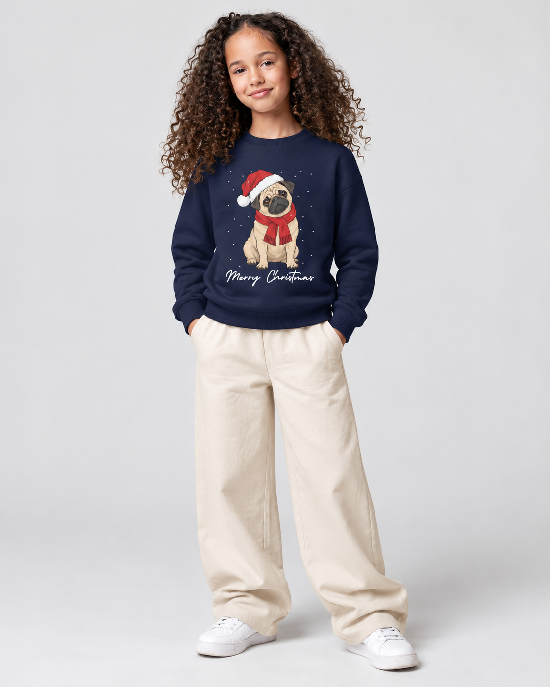 A young girl with curly brown hair stands against a light grey studio background wearing a navy blue crewneck sweatshirt with a Christmas pug graphic, cream wide-leg pants, and white sneakers.