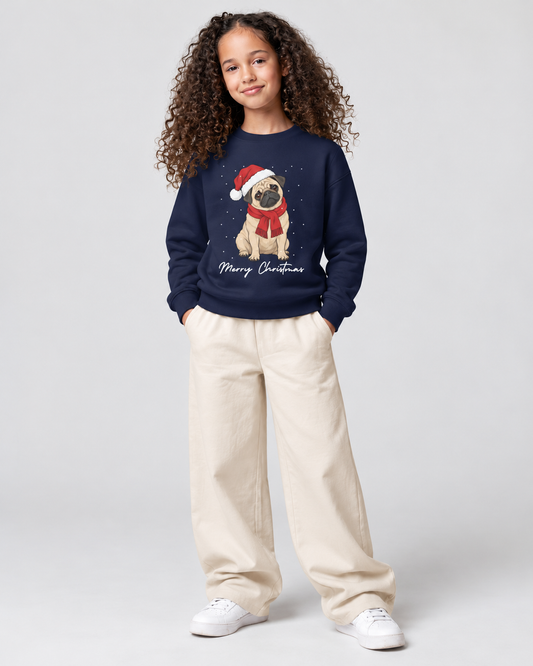 A young girl with curly brown hair stands against a light grey studio background wearing a navy blue crewneck sweatshirt with a Christmas pug graphic, cream wide-leg pants, and white sneakers.