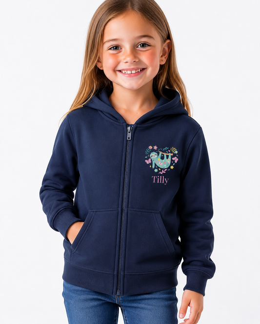A young girl with long light brown hair smiling, wearing a navy blue zip-up hoodie with a colourful floral sloth graphic and the name 'Tilly' printed on the chest, paired with blue jeans, against a plain white background.