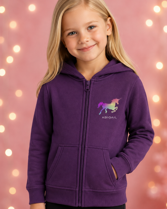 A young girl with blonde hair wearing a purple full-zip hoodie with a colorful unicorn graphic and the name 'ABIGAIL' printed on the chest, set against a pink background with warm bokeh lights.