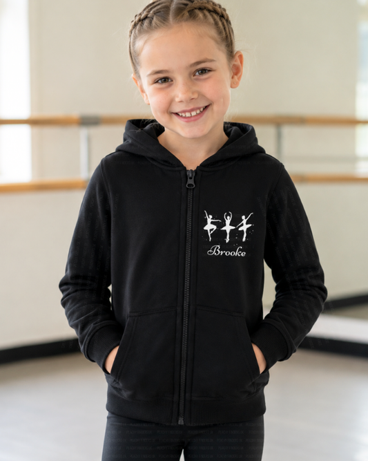 A young girl wearing a black zip hoodie featuring three white ballerina silhouettes and a personalised name print on the front - dance studio background.