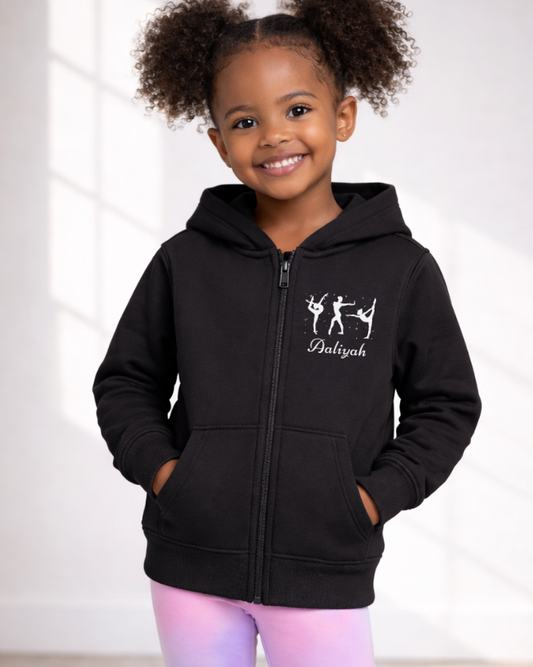 Smiling girl modelling black personalised gymnastics zip hoodie with name detail, studio portrait.