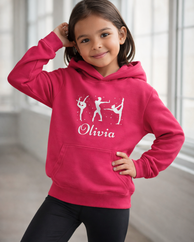 Girl wearing hot pink personalised gymnastics hoodie with white gymnast silhouettes and the name Olivia, standing in a bright studio.