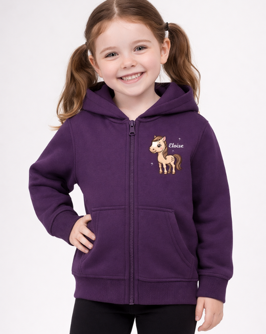 Smiling young girl wearing a personalised purple zip hoodie with cute pony graphic and name Eloise, paired with black leggings against a light grey studio background.