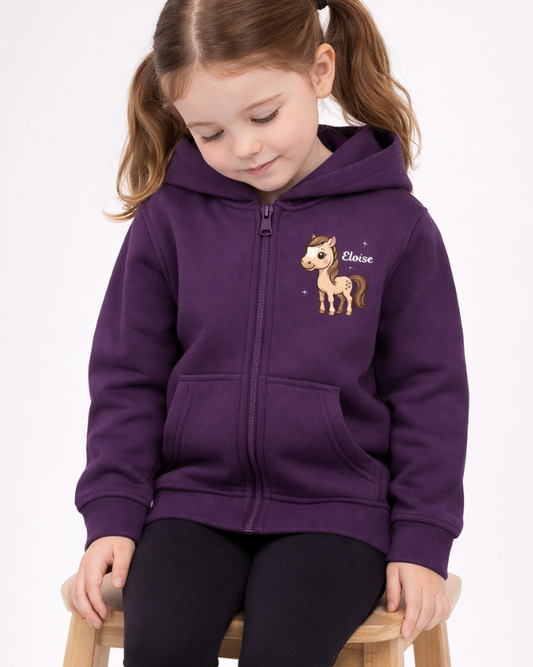 Young girl sitting on a wooden stool wearing a personalised purple zip hoodie with pony design and name Eloise, styled with black leggings on light grey studio background.
