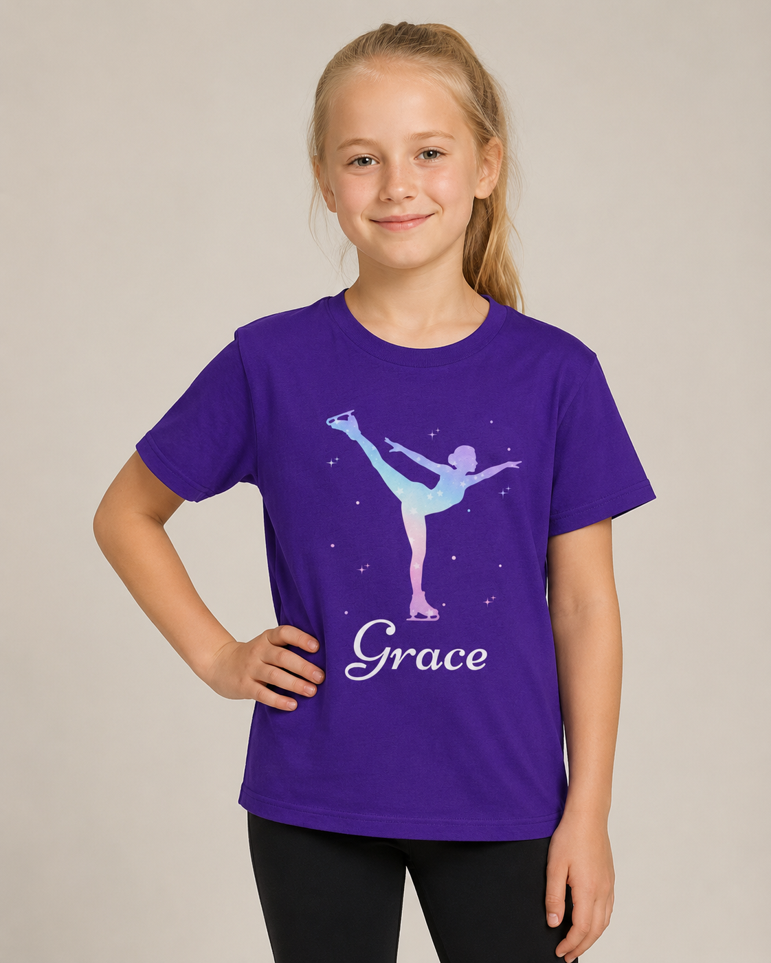 A young girl with blonde hair tied back wears a purple short-sleeve t-shirt featuring a light blue and purple ice dancer graphic with the name 'Grace' in white script. She is posing against a solid light grey studio background.
