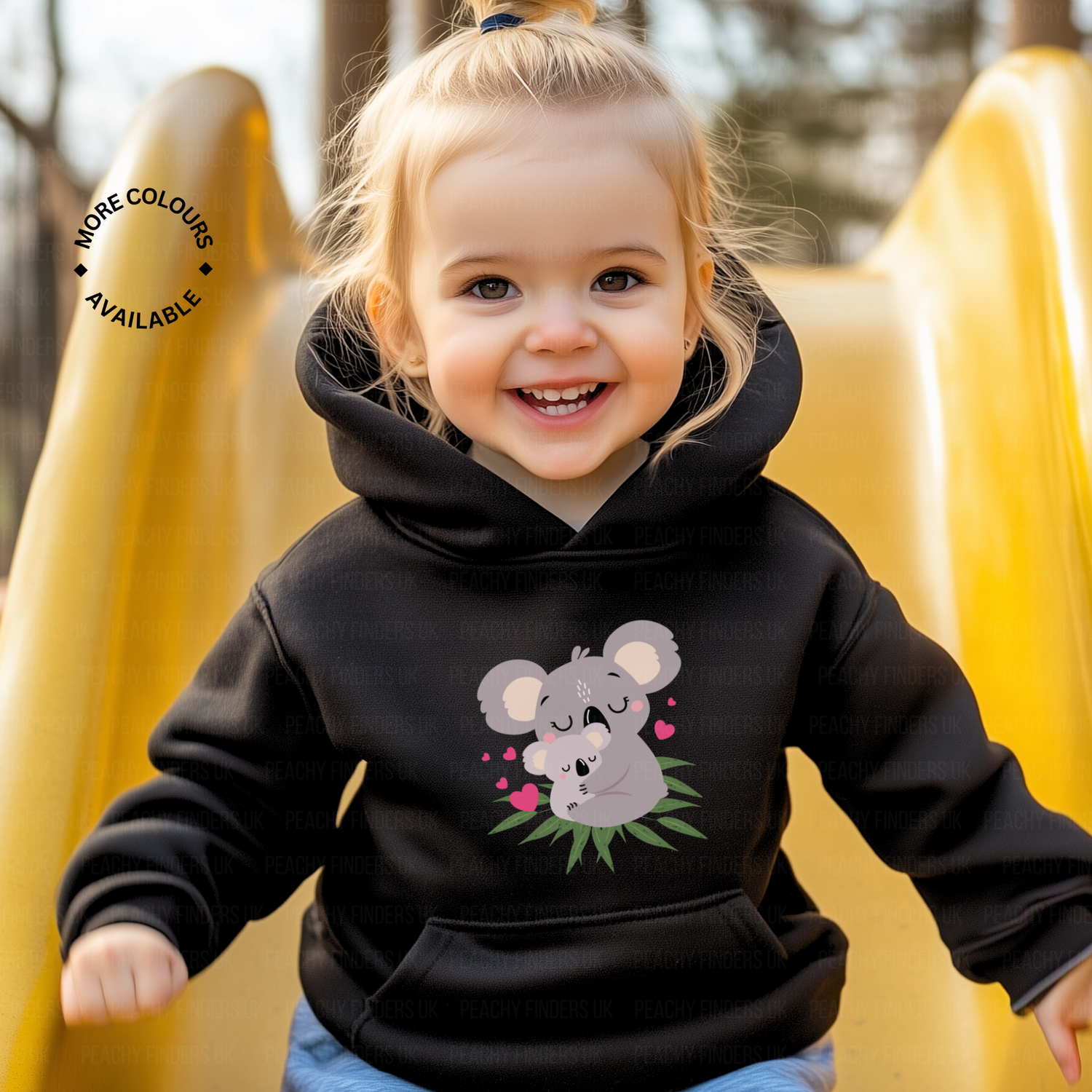 A happy toddler with blonde hair pulled up in a small ponytail smiles while sitting at the top of a yellow slide. The child wears a black hooded sweatshirt with a cute koala and baby koala graphic surrounded by pink hearts and green leaves.