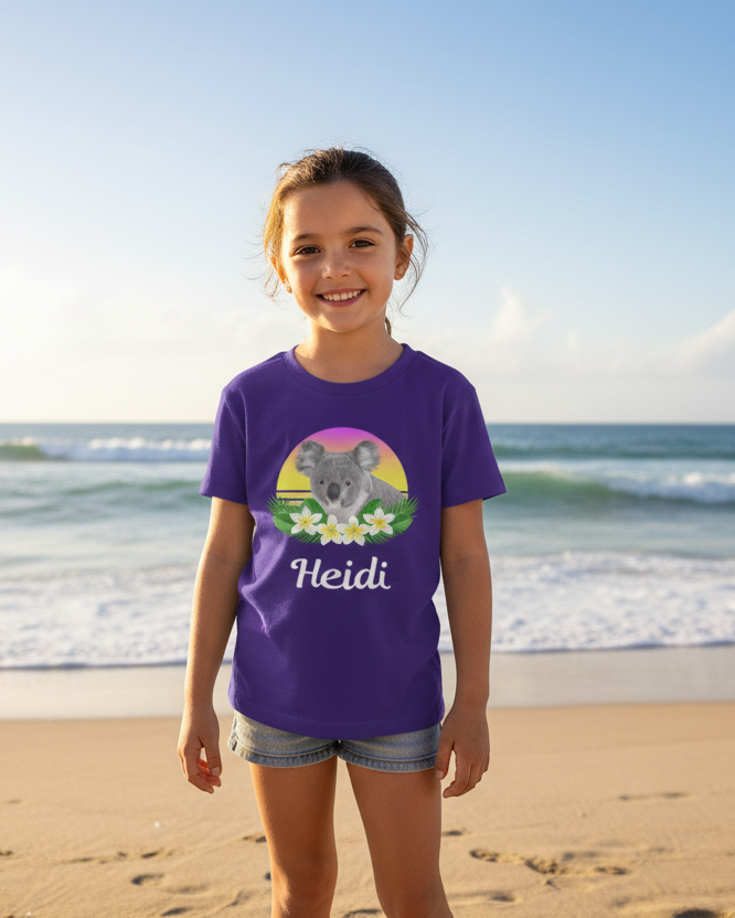 Young girl wearing a purple personalised koala t-shirt with the name Heidi, standing on a sunny beach.”