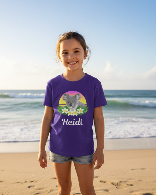 Young girl wearing a purple personalised koala t-shirt with the name Heidi, standing on a sunny beach.”