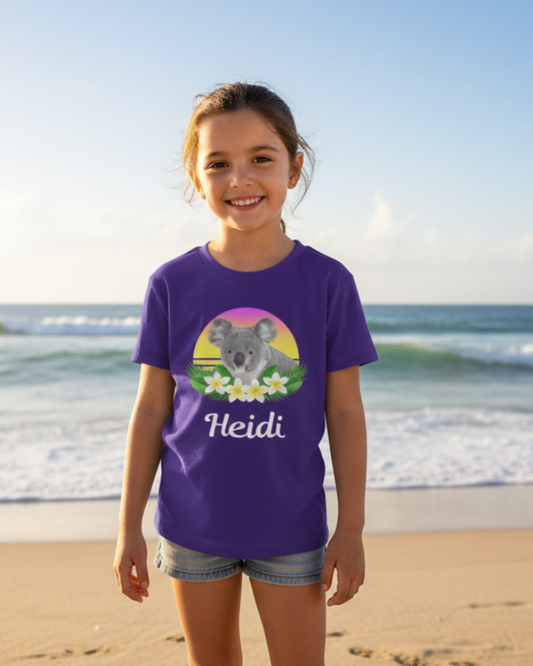 Young girl wearing a purple personalised koala t-shirt with the name Heidi, standing on a sunny beach.”