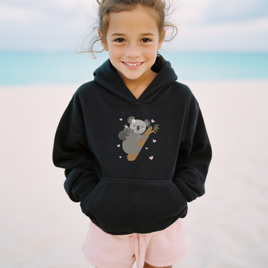 Young girl on a beach wearing a black hooded sweatshirt featuring an adorable illustration of a sleeping mother koala cuddling her baby on a tree branch, surrounded by small pink hearts.