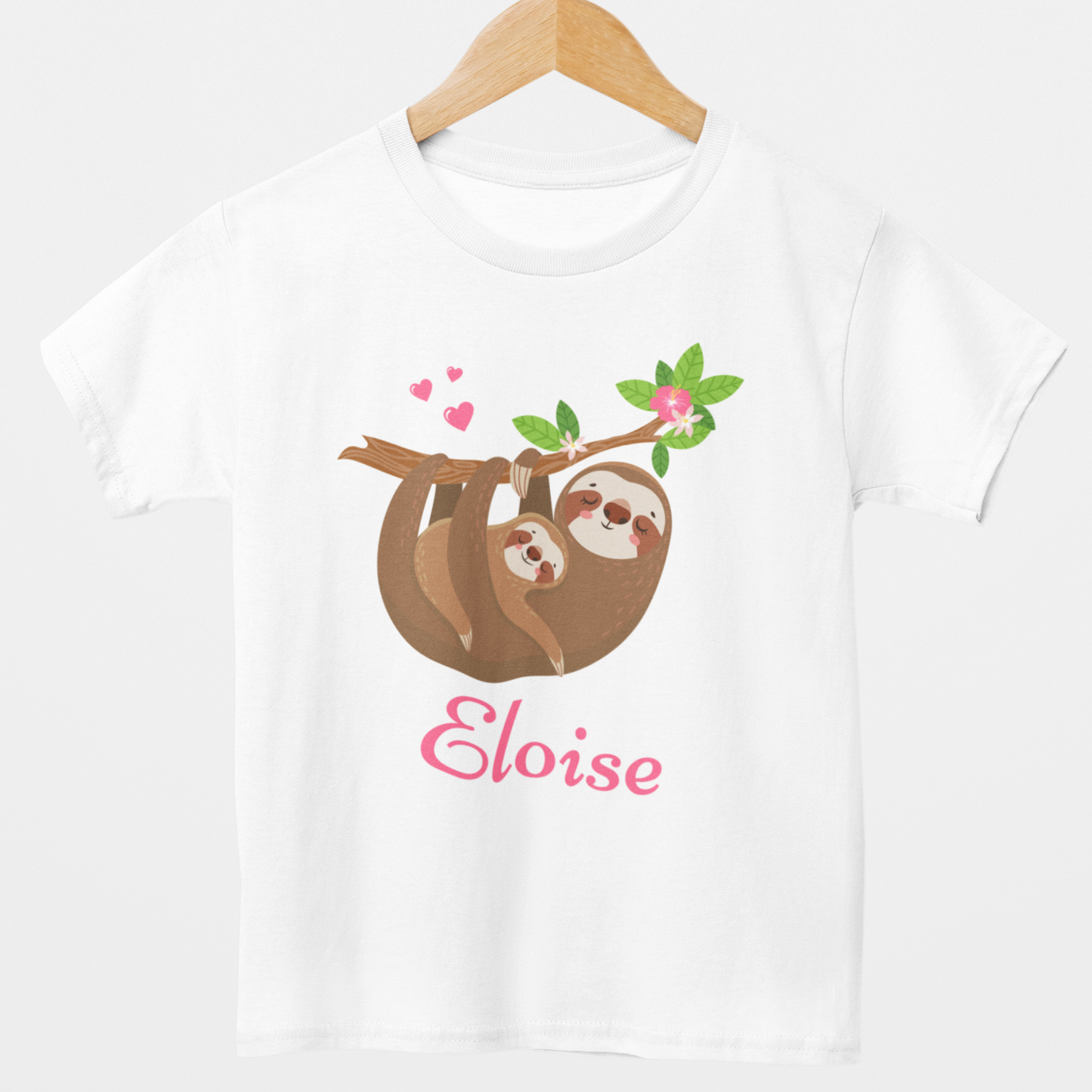 Kids T-shirt in white on a wooden hanger with a cute mum and baby sloth hanging from a leafy branch with pink hearts and flowers, and the name “Eloise” printed in pink script underneath.