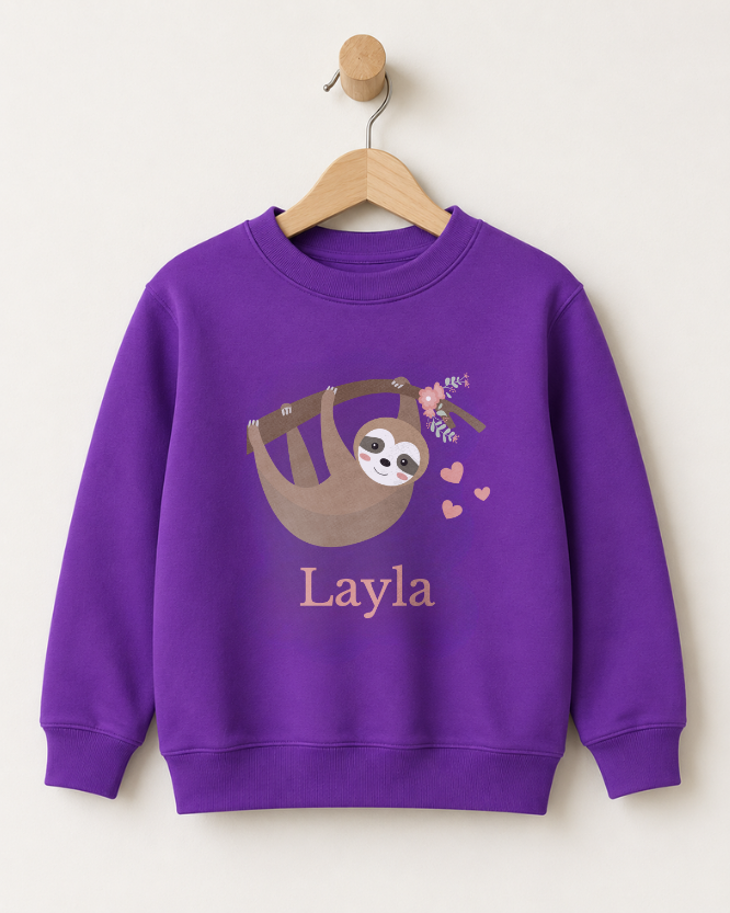 A girls purple crewneck sweatshirt hanging on a wooden hanger against a light beige wall. The sweatshirt features a cartoon sloth graphic with pink hearts and the name 'Layla' printed below in a matching pink font.