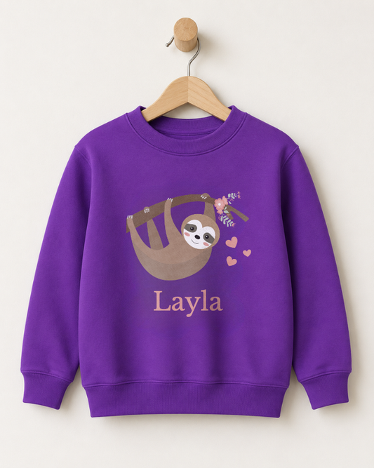 A girls purple crewneck sweatshirt hanging on a wooden hanger against a light beige wall. The sweatshirt features a cartoon sloth graphic with pink hearts and the name 'Layla' printed below in a matching pink font.