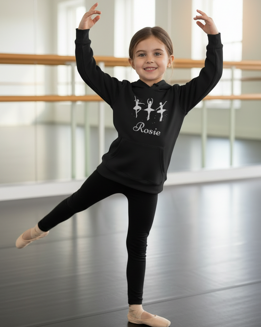 Girl in a ballet studio wearing a black personalised ballerina hoodie with the name “Rosie”, posing with arms raised and one leg extended.