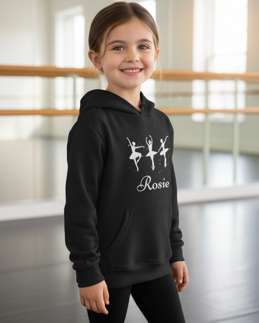 Smiling girl wearing a black personalised ballerina hoodie with “Rosie” in a ballet studio, standing relaxed with mirrors and barre softly blurred behind.