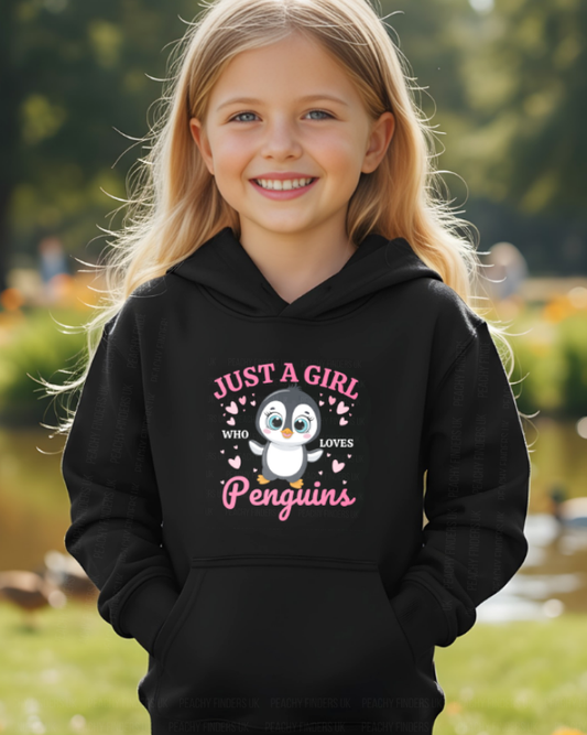 Young girl wearing a black hoodie with a printed penguin design, pink text and hearts, standing outdoors by a pond.
