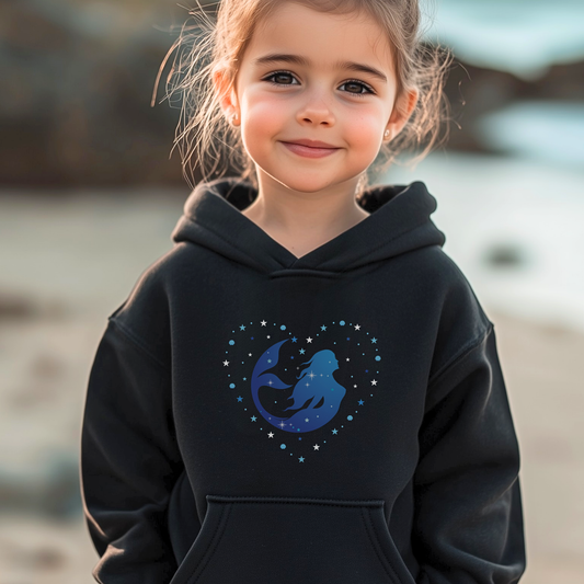 Young girl standing on a beach wearing a black hoodie featuring a blue mermaid silhouette surrounded by stars and shells in a heart shape.
