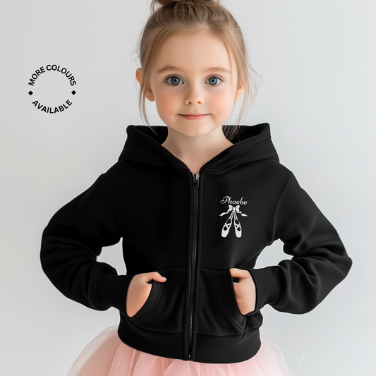 Young girl in a black personalised ballet zip hoodie with her name "Phoebe" printed above a pair of ballet shoes design, standing in a pink tutu