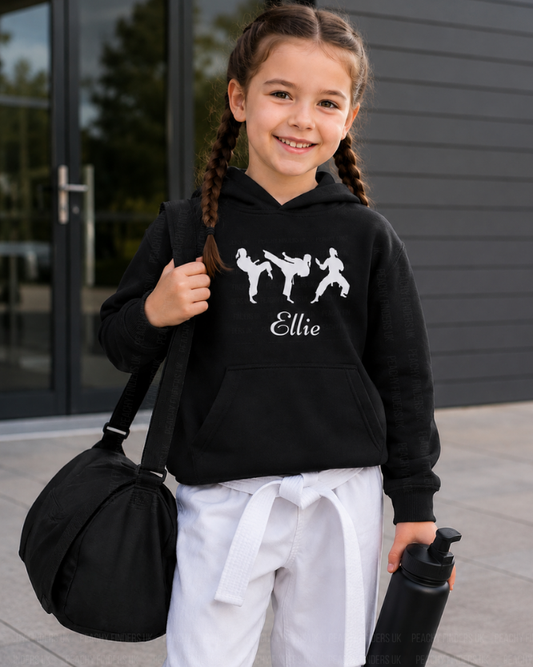 Young girl in a black personalised-martial arts/karate hoodie, holding a black bag and water bottle, standing outdoors.