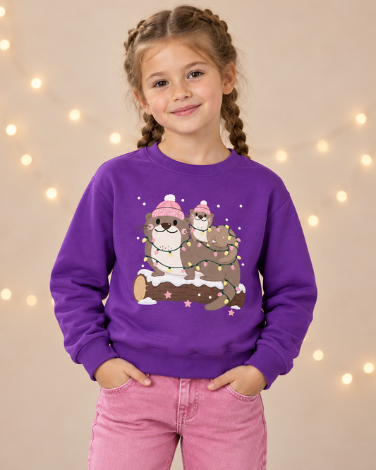 Young girl smiling while wearing a purple crewneck sweatshirt featuring two cute otters in pink bobble hats wrapped in festive fairy lights sitting on a snowy log.
