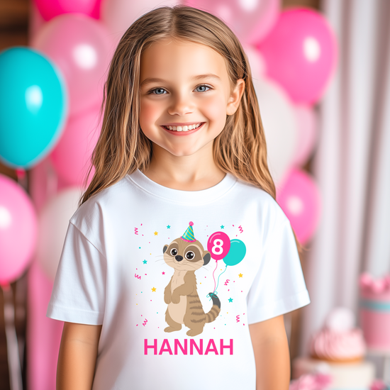 A little girl wears a white t-shirt with a meerkat wearing a party hat and balloons tied to the tail, falling coloured confetti. Personalised name in below. Pink and teal balloons in the background.