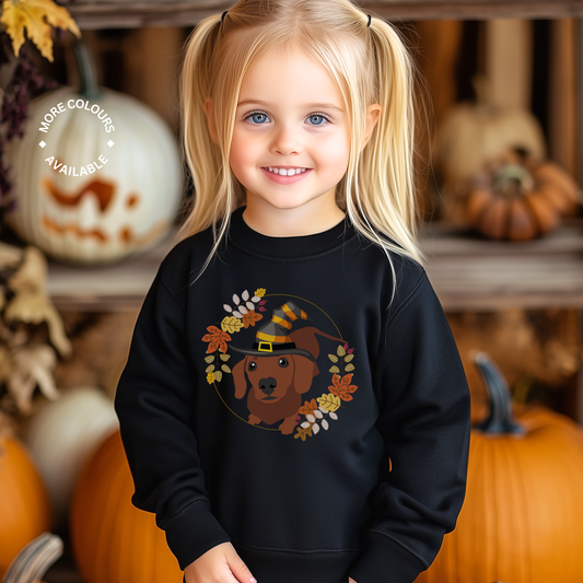 Young girl wearing black autumn sweatshirt with cute dachshund in a witch hat surrounded by fall leaves, standing in front of pumpkins