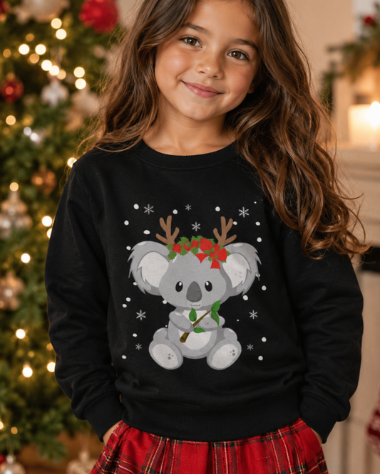 Young girl wearing a black Christmas sweatshirt with a grey koala, reindeer antlers, red festive flowers, green leaves and white snowflakes, styled with a red tartan skirt beside a softly lit Christmas tree.