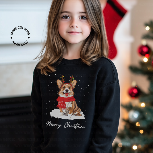 A young girl wearing a black Christmas-themed sweatshirt with a fun Corgi wearing antlers and a red festive scarf with falling snow in front of a decorated Christmas tree.
