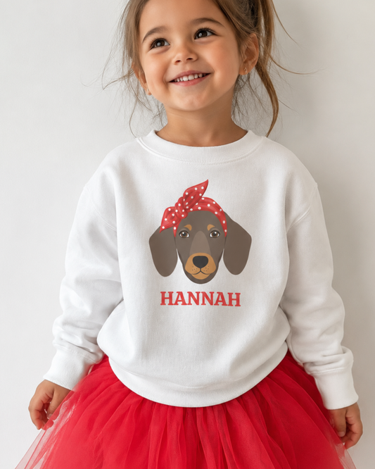A young girl smiling and wearing a white crewneck sweatshirt with a custom dachshund graphic and the name 'HANNAH' in red, paired with a red tulle tutu skirt, against a plain light background.