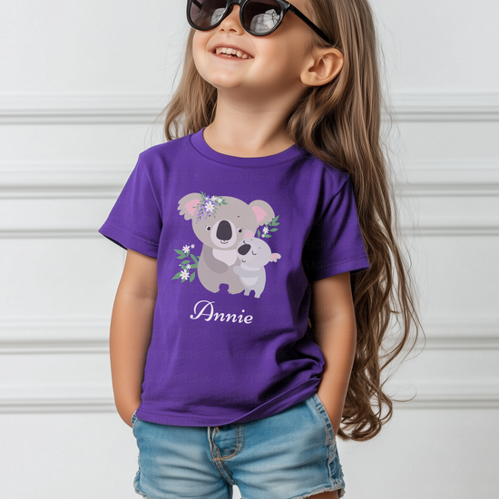 A young girl with long, light brown hair smiles and looks up and to the right while wearing sunglasses and a purple t-shirt with a koala bear design and the name 