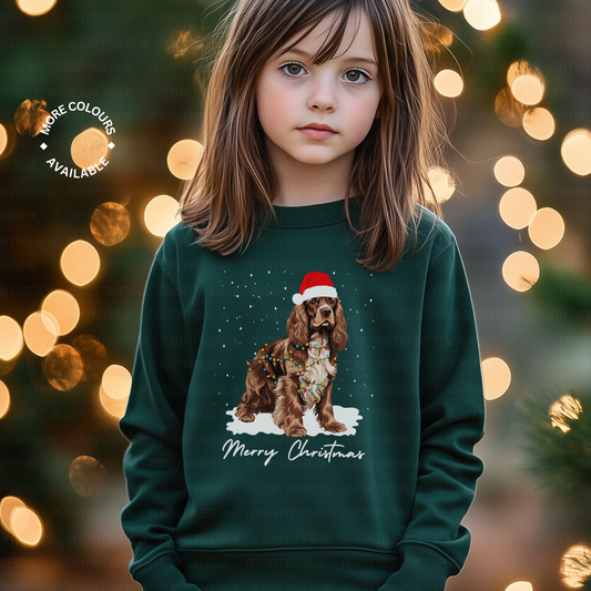 A little girl wearing a dark green Christmas sweatshirt with a brown and white Cocker Spaniel, wearing Santa hat, wrapped in Xmas lights with falling snow design.