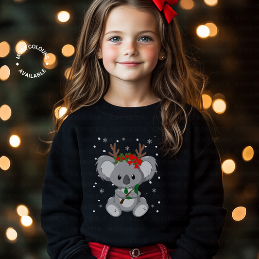 Young girl modelling a Christmas sweatshirt featuring a cute koala wearing reindeer antlers and holly, surrounded by snowflakes, printed on the front. Festive lights in the background.
